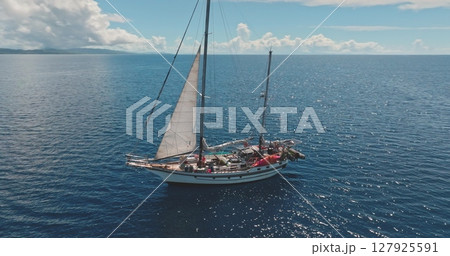 Aerial drone footage of a ketch sailing near a tropical island on a sunny day, with white sails and a wooden deck, surrounded by blue waters and a distant coastline. Wild sea nature travel background 127925591