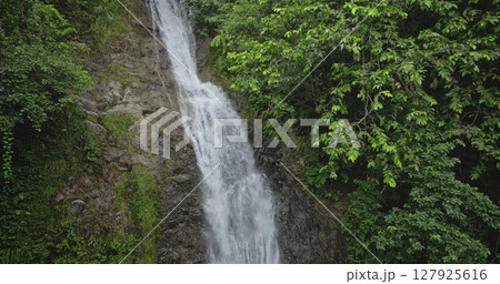 Refreshing waterfall cascading down rocky cliffs, surrounded by vibrant green vegetation, picturesque scene in tropical rainforest, captured with aerial drone footage. Amazing nature travel background 127925616