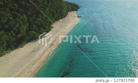 Capturing aerial drone footage of a tropical beach merging with turquoise waters on a sunny day, surrounded by lush green forests covering the nearby hills and coastline Capturing aerial drone footage of a tropical beach merging with turquoise waters on a sunny day, surrounded by lush green forests covering the nearby hills and coastline 127925700