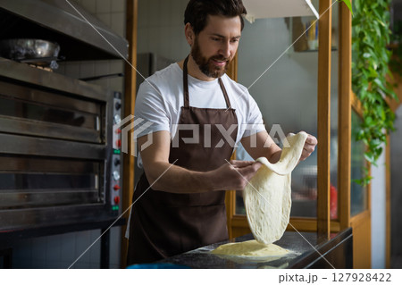 Bearded man wearing a white t-shirt and brown apron tosses pizza dough in a bakery kitchen Bearded man wearing a white t-shirt and brown apron tosses pizza dough in a bakery kitchen 127928422