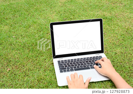 Hands typing on laptop with blank screen placed directly on the grass 127928873