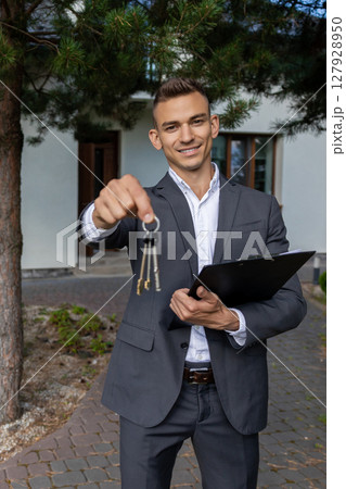Real estate agent presenting house keys and documents on clipboard in front of property for sale 127928950