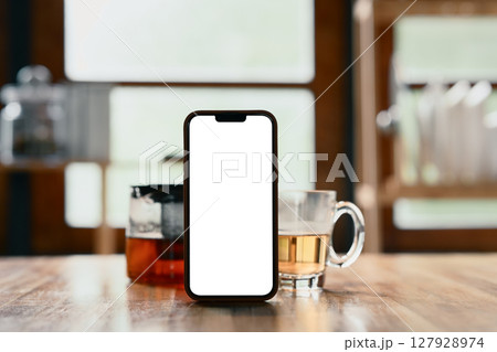 Blank screen smartphone placed on a wooden kitchen counter with hot tea 127928974