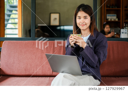 Smiling woman enjoying coffee while working on a laptop in a cozy, modern living room environment. 127929967