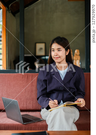 A woman in a blazer sits on a couch with a laptop and notebook, embodying remote work in a modern home office environment. A woman in a blazer sits on a couch with a laptop and notebook, embodying remote work in a modern home office environment. 127929970