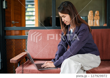 A focused young woman using a laptop while sitting on a comfortable sofa in a stylish home office environment. A focused young woman using a laptop while sitting on a comfortable sofa in a stylish home office environment. 127929972