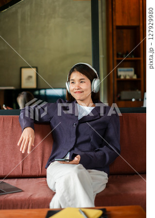Woman enjoying music with headphones, sitting on a sofa in a stylish living room, exuding relaxation and comfort. 127929980