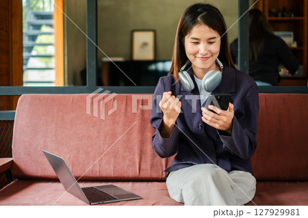 A young woman with headphones celebrates success while using her smartphone and laptop in a modern home office. A young woman with headphones celebrates success while using her smartphone and laptop in a modern home office. 127929981