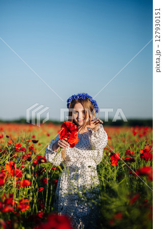 Young woman with blue flowers crown holding a bouquet of red poppies and gesturing in a field of papaver rhoeas Young woman with blue flowers crown holding a bouquet of red poppies and gesturing in a field of papaver rhoeas 127930151