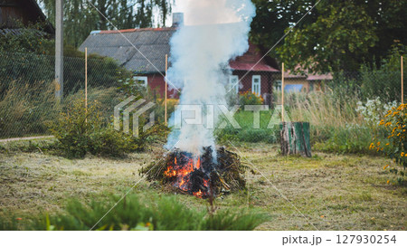 Smoldering tree branches creating dense smoke plume near wooden house, rural garden setting with rustic wooden fence 127930254