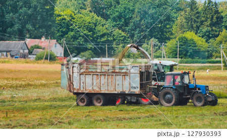 Rural tractor unloading golden hay bales into trailer, sunlight illuminating agricultural landscape near traditional village Rural tractor unloading golden hay bales into trailer, sunlight illuminating agricultural landscape near traditional village 127930393