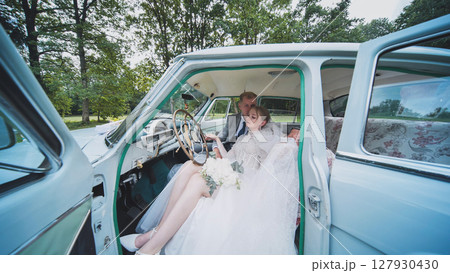 Newlyweds embracing inside classic automobile, sharing intimate moment during wedding celebration with vintage automotive backdrop 127930430