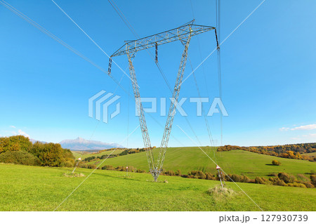 Large metal power pylon under electricity lines built in countryside with green grass covered hills and small forests, mount Krivan (Slovak symbol) at distance, blue sky background Large metal power pylon under electricity lines built in countryside with green grass covered hills and small forests, mount Krivan (Slovak symbol) at distance, blue sky background 127930739