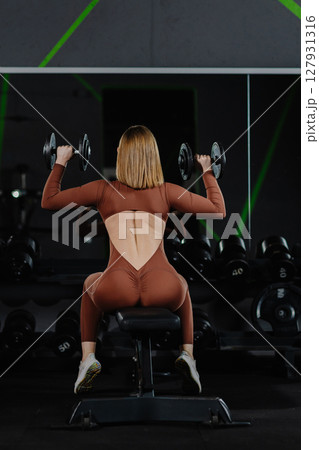 Determined sportswoman lifting dumbbells while sitting on bench in gym Determined sportswoman lifting dumbbells while sitting on bench in gym 127931316