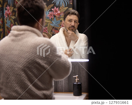 Middle aged man in bathrobe grooming his beard with a brush in bathroom mirror reflection 127931661