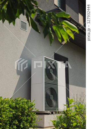 Vertical outdoor heat pump unit installed next to a house wall, surrounded by green shrubs in a modern landscaped garden Vertical outdoor heat pump unit installed next to a house wall, surrounded by green shrubs in a modern landscaped garden 127932444