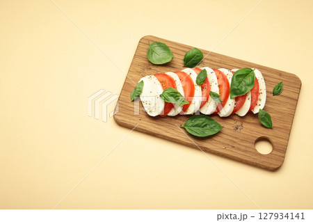 Mozzarella cheese with green basil leaves and tomato on cutting board on beige background 127934141