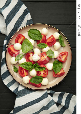 Italian Caprese salad with cherry tomatoes, mozzarella, basil on plate on black background. Vertical photo 127934150