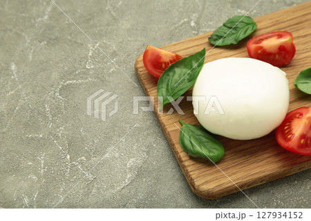 Mozzarella cheese with green basil leaves and tomato on cutting board on grey background 127934152