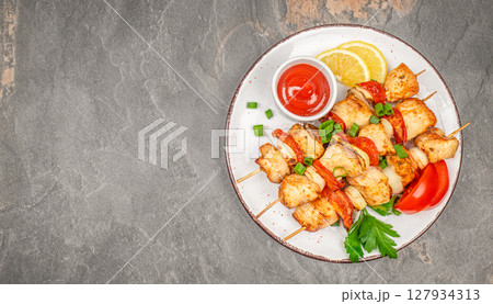 Grilled chicken skewers served with fresh vegetables, herbs, and tomato sauce on a rustic plate over a dark stone background 127934313