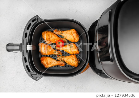 Top view of seasoned chicken drumsticks and a tomato cooking inside a modern black air fryer on a light kitchen counter Top view of seasoned chicken drumsticks and a tomato cooking inside a modern black air fryer on a light kitchen counter 127934336
