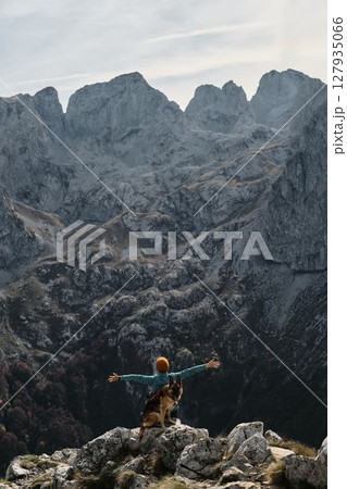 A female traveler raises her arms in triumph while standing with her German Shepherd atop a rocky ridge in Prokletije National Park, Montenegro. Rear view. Travel with pet concept 127935066