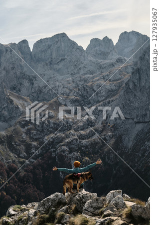 A female traveler raises her arms in triumph while standing with her German Shepherd atop a rocky ridge in Prokletije National Park, Montenegro. Rear view. Travel with pet concept 127935067
