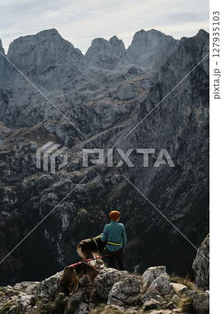 A woman stands on a rocky cliff with her German and Australian Shepherds, overlooking the dramatic peaks of Prokletije National Park in autumn, Montenegro. Hiking with pets concept A woman stands on a rocky cliff with her German and Australian Shepherds, overlooking the dramatic peaks of Prokletije National Park in autumn, Montenegro. Hiking with pets concept 127935103