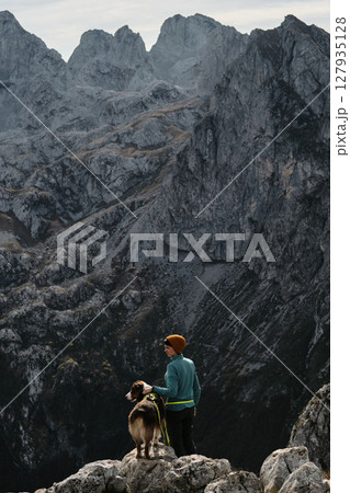 A woman and her Australian shepherd dog enjoy the majestic mountain view from a rocky ridge in Prokletije National Park, Montenegro, during fall. Rear view. Hiking with pet concept 127935128