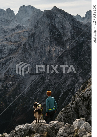 A woman and her Australian shepherd dog enjoy the majestic mountain view from a rocky ridge in Prokletije National Park, Montenegro, during fall. Rear view. Hiking with pet concept 127935150