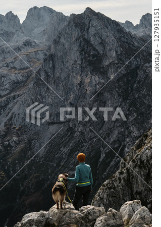 A woman and her Australian shepherd dog enjoy the majestic mountain view from a rocky ridge in Prokletije National Park, Montenegro, during fall. Rear view. Hiking with pet concept 127935151