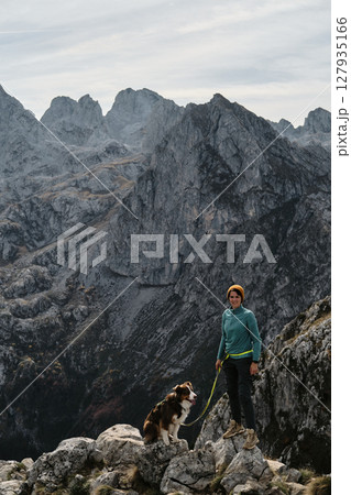 A female hiker stands proudly on a rocky cliff with her Australian Shepherd dog, framed by the dramatic peaks of Prokletije, Montenegro in autumn season. Hiking with pet concept 127935166