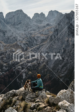 Young woman sits on a rocky summit beside her Australian Shepherd dog, admiring the rugged mountains of Prokletije National Park in autumn Montenegro. Hiking with pet concept 127935167