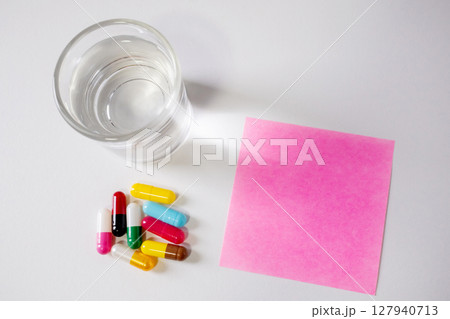 Top view of glass of water, many pill capsules and pink note paper on white background with copy space. Top view of glass of water, many pill capsules and pink note paper on white background with copy space. 127940713