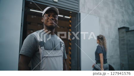 Outside of Logistics Warehouse: Worker Giving Cardboard Box to Colleague, Looking at Camera Outside of Logistics Warehouse: Worker Giving Cardboard Box to Colleague, Looking at Camera 127941576