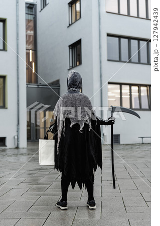 Back view of kid in grim reaper Halloween costume with scythe and bag for sweets at school building facade ready for Halloween party celebration. Child in scary spooky black death suit outdoors 127942439