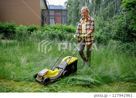 Woman using electric lawn mower in overgrown garden near wooden house 127942961