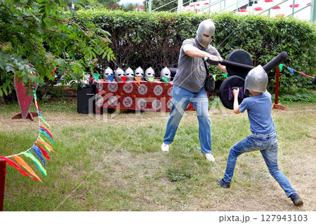 Father and child wearing helmets engage in playful medieval sword fight with foam swords and shields on summer day 127943103