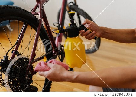 Mountain bike owner spraying and wiping down bicycle with cloth, performing detailed maintenance after intense trail riding, preserving cycling gear condition and functionality at home, close-up. 127943372