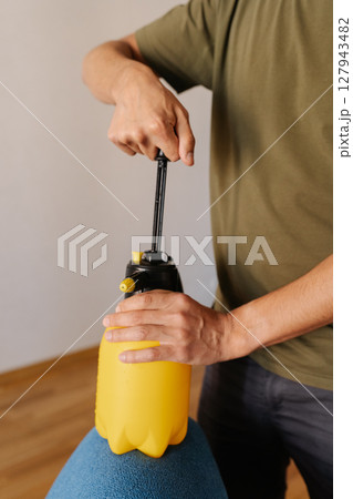Vertical cropped shot of man pumping yellow water sprayer bottle while holding on blue sofa, preparing to clean or disinfect upholstery at home. Concept of domestic work, housekeeping and household. Vertical cropped shot of man pumping yellow water sprayer bottle while holding on blue sofa, preparing to clean or disinfect upholstery at home. Concept of domestic work, housekeeping and household. 127943482