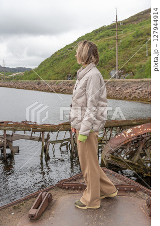 Woman in beige jacket and sunglasses standing by a calm river with an old wooden shipwreck and winding coastal road in the background 127944914