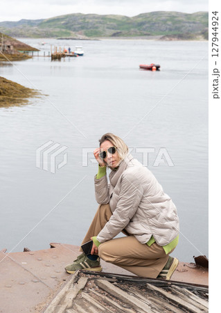 Woman in beige jacket and sunglasses crouching thoughtfully by the water with distant boats and green hills in the background 127944924