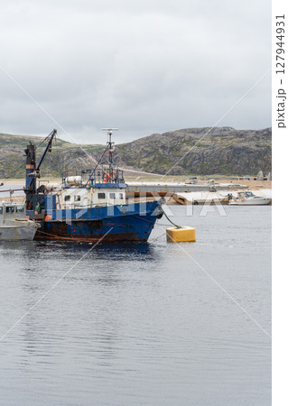 Old blue fishing vessel anchored near the shore with rugged hills in the background and cloudy sky above 127944931