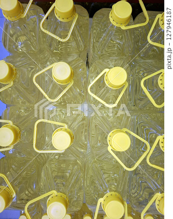 Large plastic bottles filled with vegetable sunflower oil are neatly organized on store shelves. The yellow caps stand out against the transparent packaging, highlighting the product. 127946187