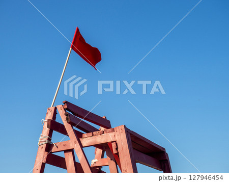 Red flag on a wooden platform. Flag as a symbol of danger. Bathing is prohibited. Beach on the sea. The concept of rescue work. 127946454