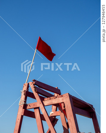 Red flag on a wooden platform. Flag as a symbol of danger. Bathing is prohibited. Beach on the sea. The concept of rescue work. Red flag on a wooden platform. Flag as a symbol of danger. Bathing is prohibited. Beach on the sea. The concept of rescue work. 127946455