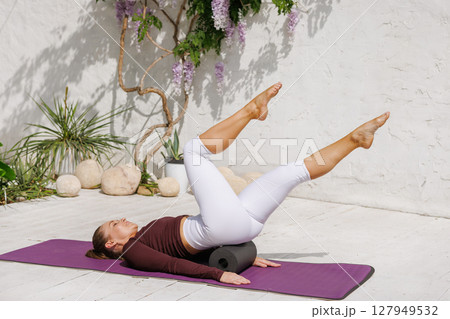 Athletic woman performing pilates with foam massage roller for hip and back recovery on yoga mat in bright studio 127949532