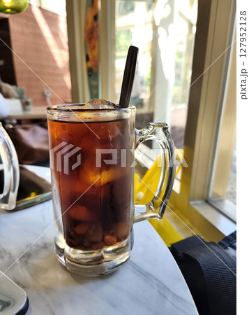 Lo Han Kuo or Monk Fruit Drink, an iced herbal beverage with a dark amber hue and a spoon, set by a sunlit window. 127952128