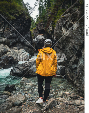 A young woman stands on rocky terrain near a flowing river in a canyon. 127953015