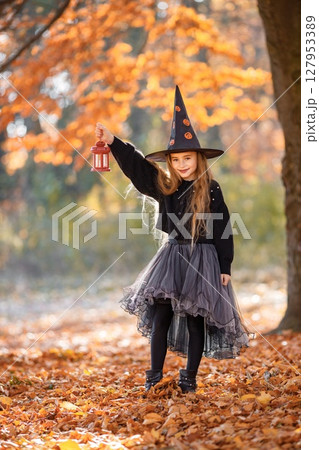 Cute happy little girl dressed in witch costume standing with over autumn forest background. Girl wearing dress and cone hat and holding a lantern. Girl dressed for Halloween. Cute happy little girl dressed in witch costume standing with over autumn forest background. Girl wearing dress and cone hat and holding a lantern. Girl dressed for Halloween. 127953389